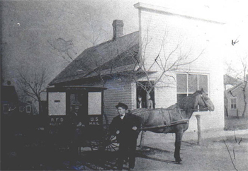 Black & white photo of man next to a carriage drawn by a horse, in front of a small building