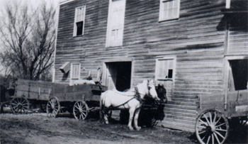 Black & white photo of a wagon drawn by two horses in front of wooden building